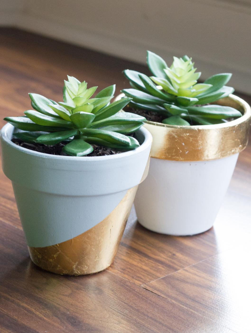 Two white painted terracotta pots with gold leaf bands and diagonals, each holding a green succulent on a wood table