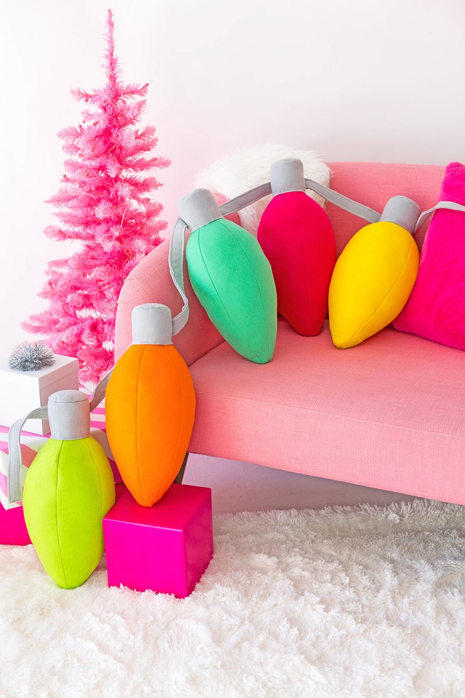 Oversized felt Christmas light bulb pillows in pink yellow and blue with gray tops piled up on a sofa