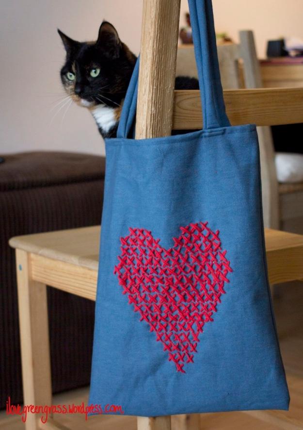 Denim blue tote bag on a chair with a large red cross stitched heart on the front and a tabby cat behind it
