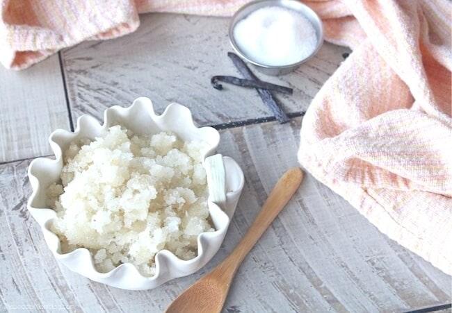 Small white scalloped dish of vanilla honey sugar lip scrub with a wooden spoon and a pink cloth on a white wood table