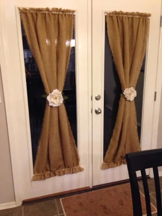 Two burlap curtain panels hanging on French doors, each tied back in the middle with a large white fabric flower