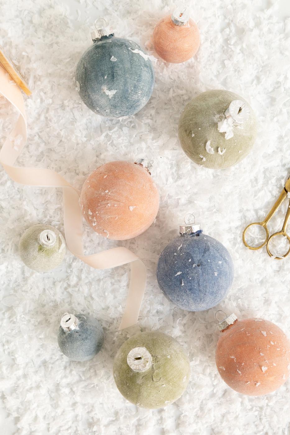 Velvet look ball ornaments in dusty blue, sage green and peach lying in fake snow with a pale pink ribbon and gold scissors