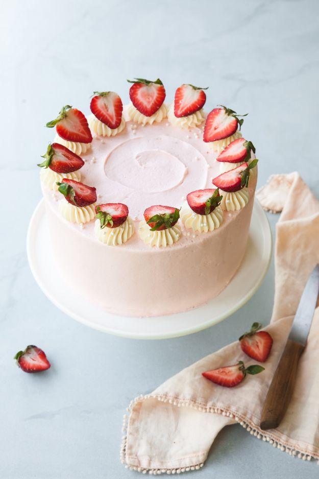 Pale pink strawberry layer cake on a white stand with halved berries piped around the top edge and a knife