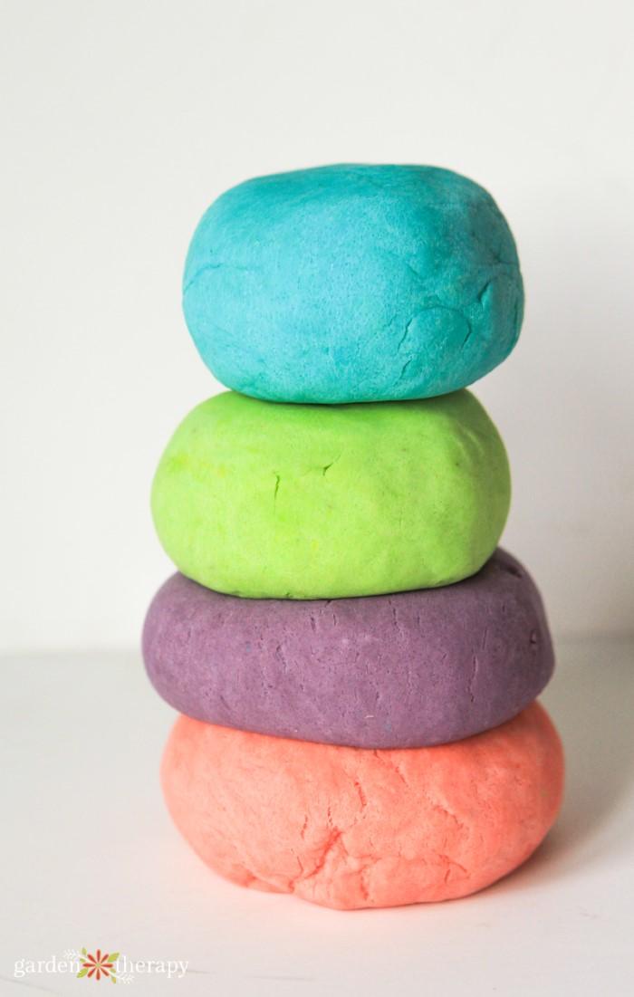 Four stacked balls of homemade playdough in bright teal, lime green, purple and coral on a white surface