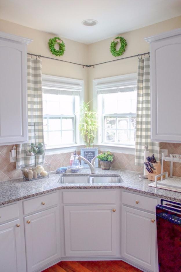 Gray and white buffalo check cafe curtains at two corner kitchen windows above a sink, with wreaths on white cabinets