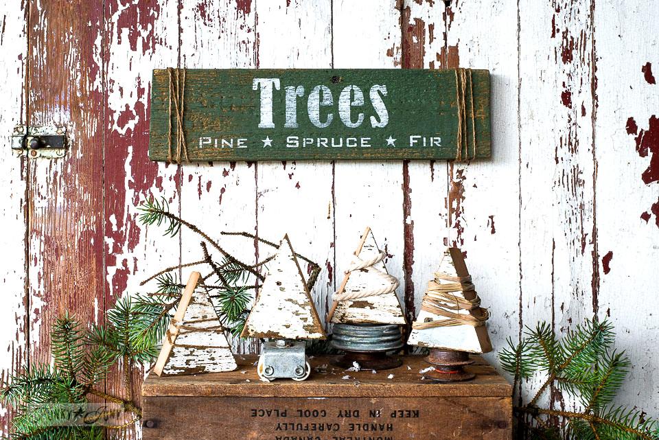 Four chippy white scrap wood trees wrapped in twine and rope on rusty junk stands under a green stenciled Trees sign