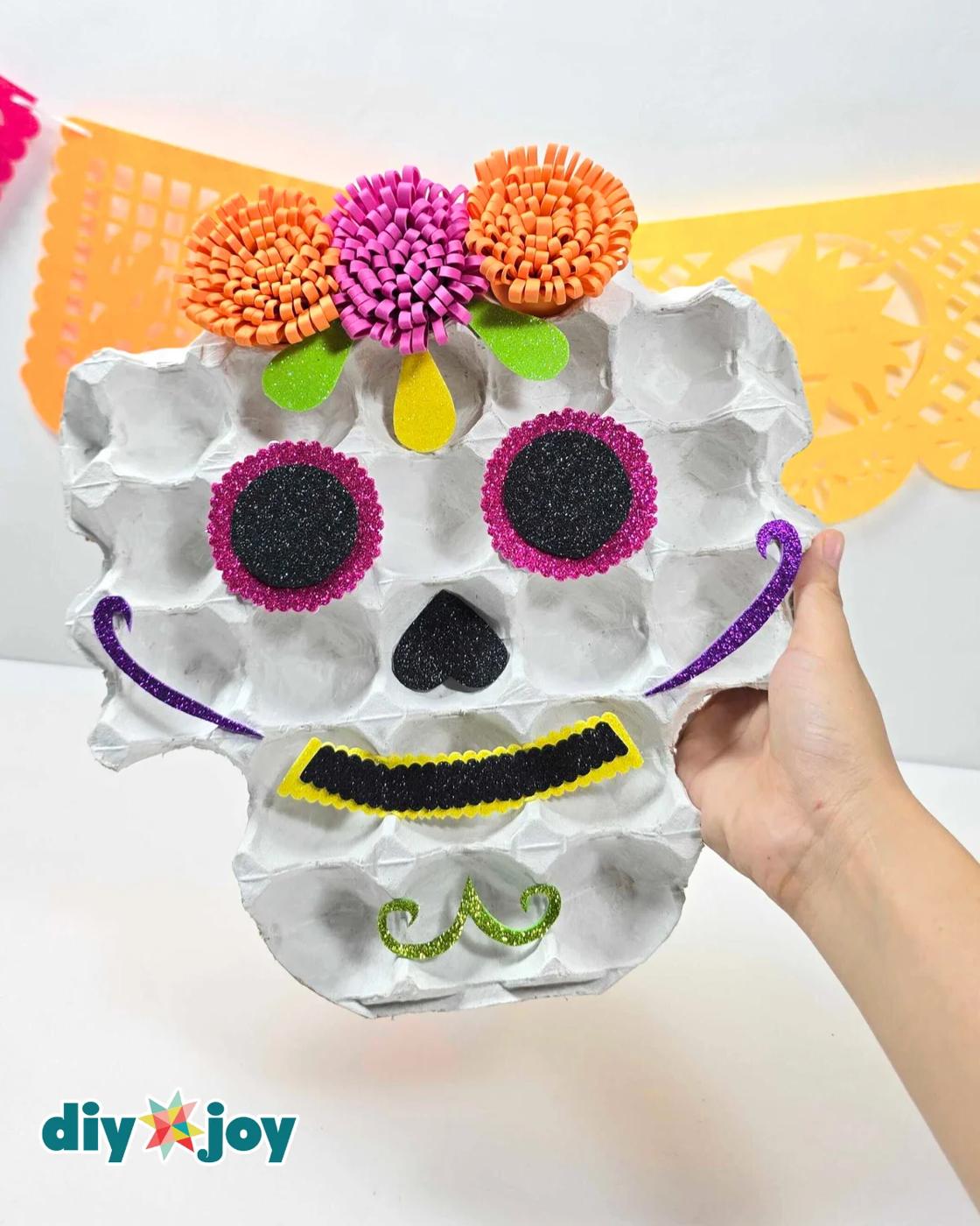 Dia de los Muertos decor - a hand holding the finished egg tray sugar skull upright in front of an orange and pink papel picado banner