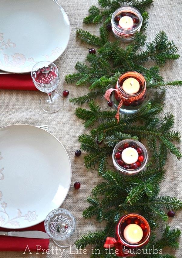 Mason jars in two sizes with water, fresh cranberries and floating candles, tied with ribbon among fresh evergreen sprigs