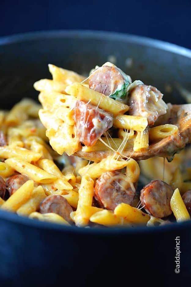 One pot penne pasta with smoked sausage and melted cheese being lifted from a dark blue pot on the stove