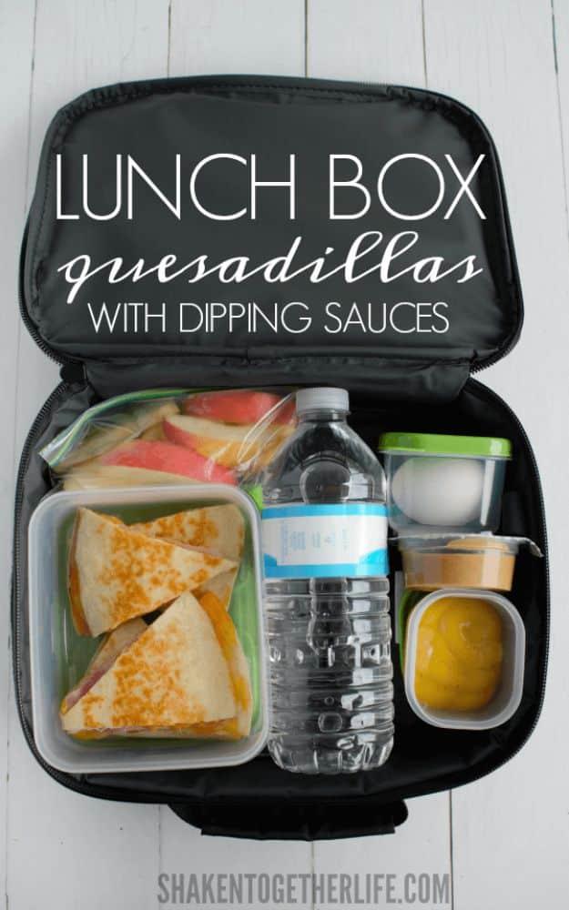 Lunch box quesadillas packed in a black bento container with small pots of dipping sauce, fruit and water