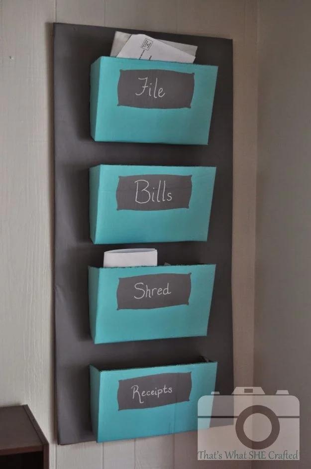 Four turquoise cardboard pockets labelled file, bills, shred and receipts on a gray painted cardboard backboard on a wall