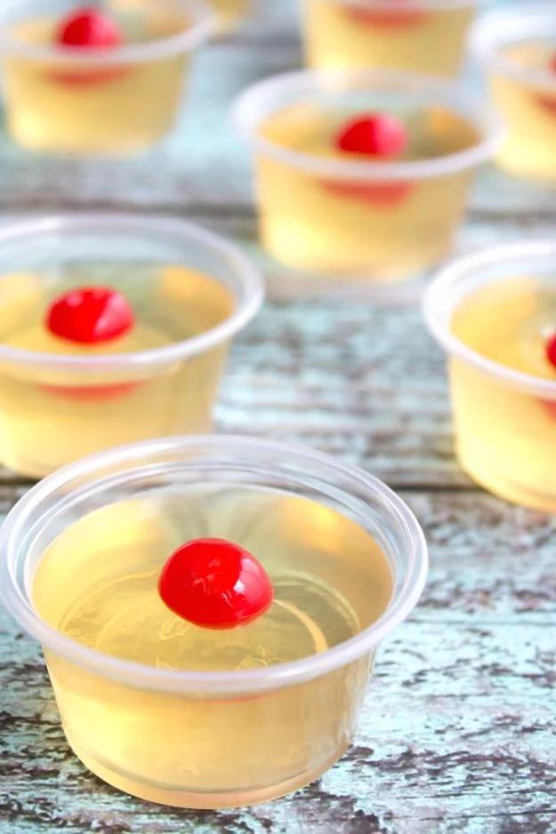 Amaretto sour jello shots in lidded two ounce plastic ramekins, amber colored with a cherry in each one