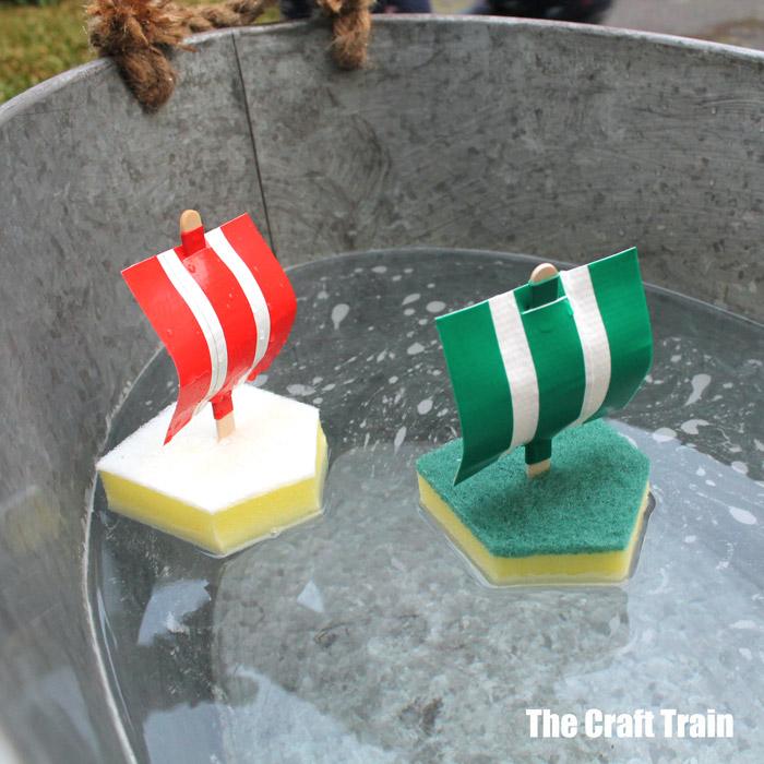 Two sponge boats with striped red and green plastic sails on craft stick masts floating in a galvanized tub of water
