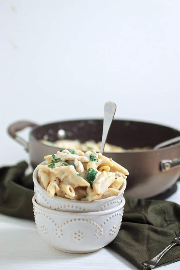 Chicken spinach alfredo penne in a small white bowl with a skillet of creamy sauce in the background