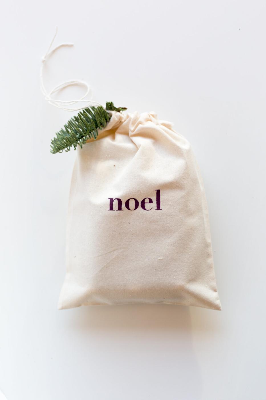 A natural cotton drawstring gift bag stenciled with the word noel in deep purple, tied with a fir sprig