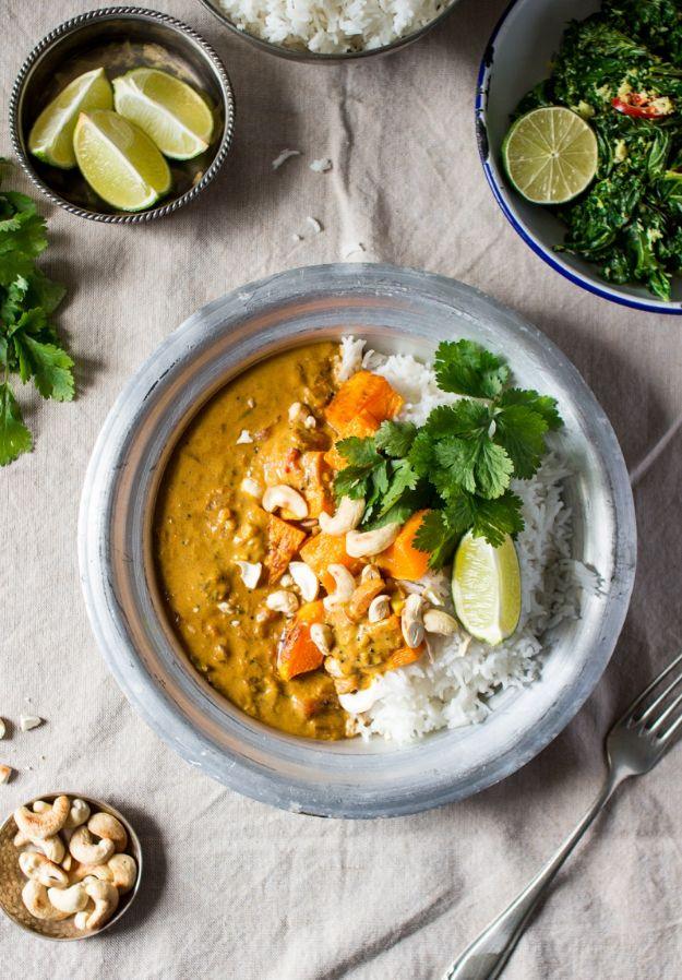 Metal bowl of pumpkin coconut curry with white rice, cashews, cilantro and lime wedges on a rustic table