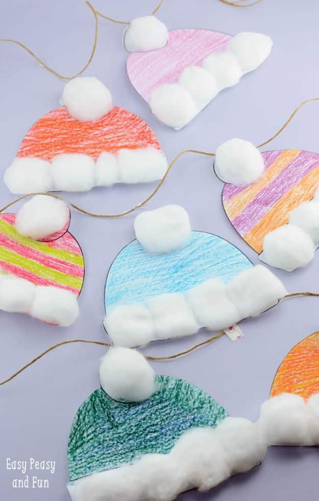 Six paper winter hats colored in crayon with cotton ball brims and pom poms, strung across a pale lavender background