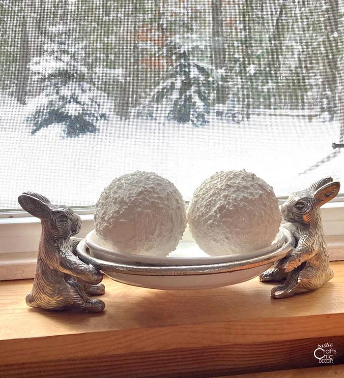 Two sparkly white faux snowballs sitting in a silver dish with rabbit handles on a windowsill in front of snowy woods