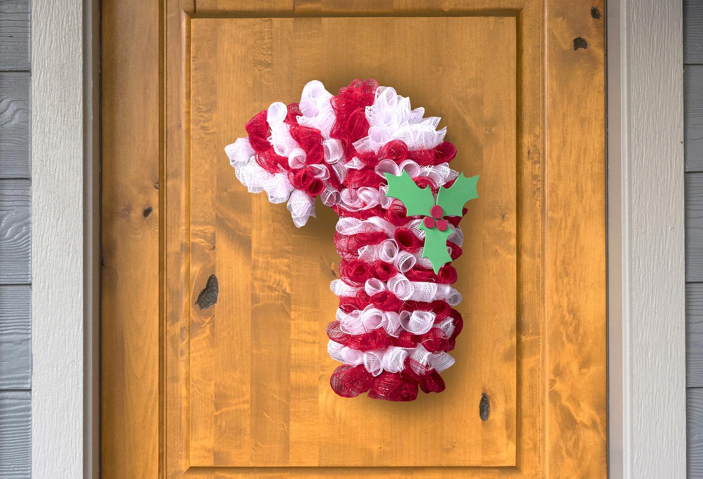 A candy cane shaped wreath of curled red and white deco mesh with green holly leaves and red berries hanging on a wooden door