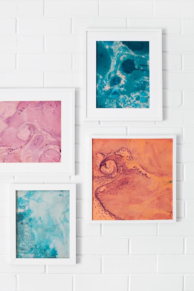 Four white framed marbled paper prints in teal, pink, orange and aqua hung at staggered heights on a white brick wall