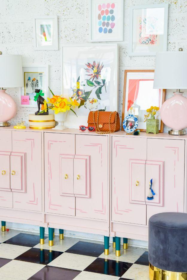 Three pale pink cabinets with raised panel doors on green and brass legs, gallery wall above and black and white tile floor
