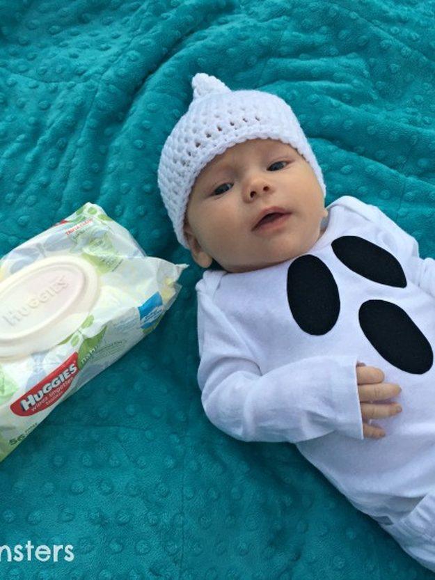 Baby wearing a white ghost bodysuit with black face shapes and a pointed crocheted hat on a teal blanket