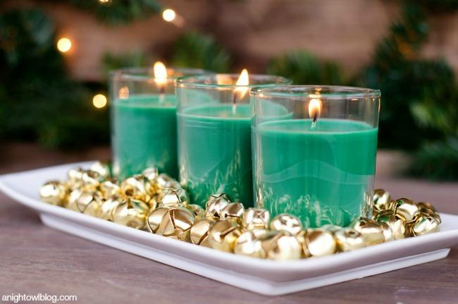 Three green scented jar candles on a white platter surrounded by gold jingle bells in two sizes on a dark wood table