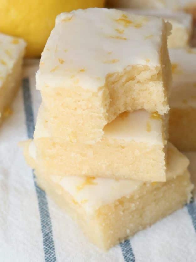 Glazed lemon brownies stacked on a striped cloth with a pale yellow crumb and white lemon glaze on top