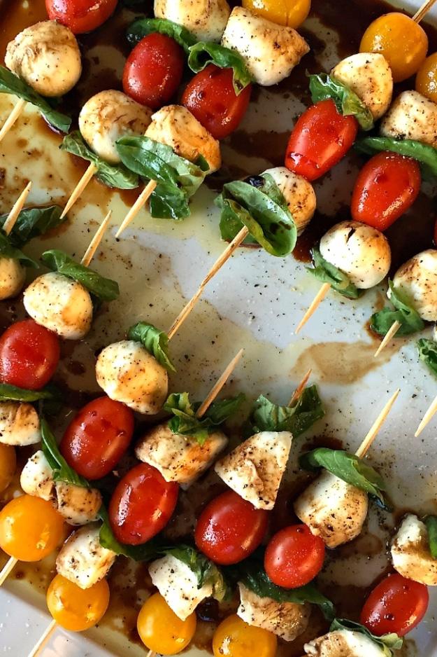 Mini caprese skewers lined up on a platter with cherry tomatoes, mozzarella balls and basil leaves beside the grill