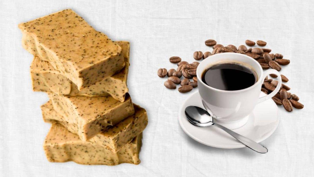 Homemade coffee soap, stacked pale soap bars flecked with coffee grounds beside a white cup of black coffee and beans
