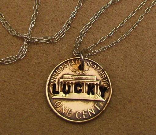 Penny pendant hammered flat and stamped with the word LUCKY, hanging on fine silver chain against warm brown