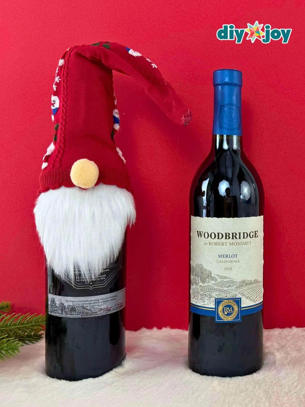 Wine bottle dressed as Santa with a red Christmas sock hat, a white faux fur beard and a pom pom nose beside a plain bottle