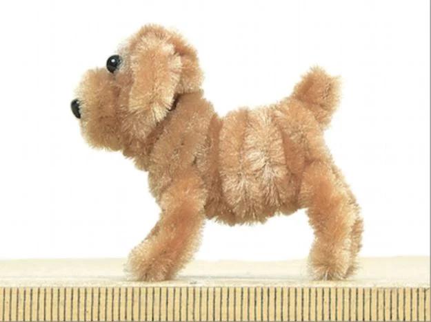 Small fluffy dog made from a single beige pipe cleaner with black toy eyes and nose, standing on a pale wooden ruler