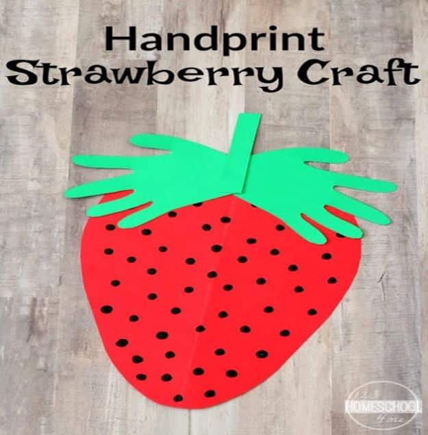 Red paper strawberry with black marker seeds topped by two green paper handprints as leaves, on a weathered wooden background