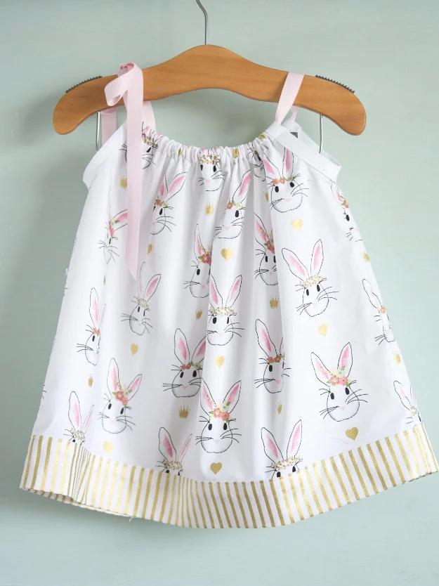White bunny print pillowcase dress with a gold striped hem band and pink ribbon ties, on a wooden hanger