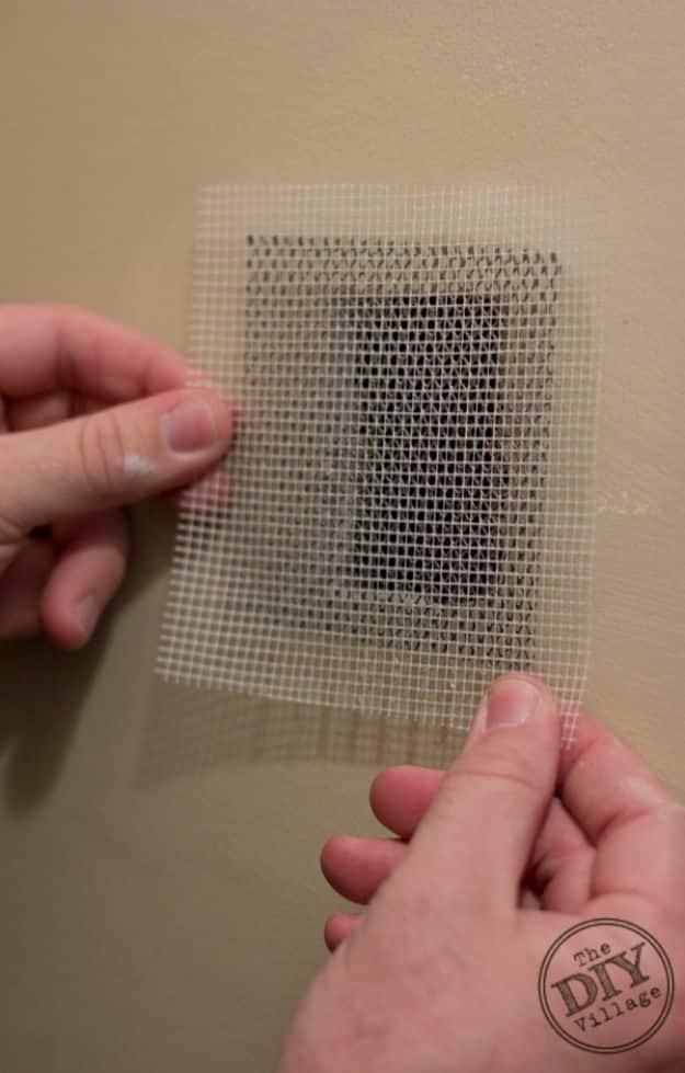 Drywall patch repair, two hands pressing a square self-adhesive mesh drywall patch over a hole on a tan painted wall
