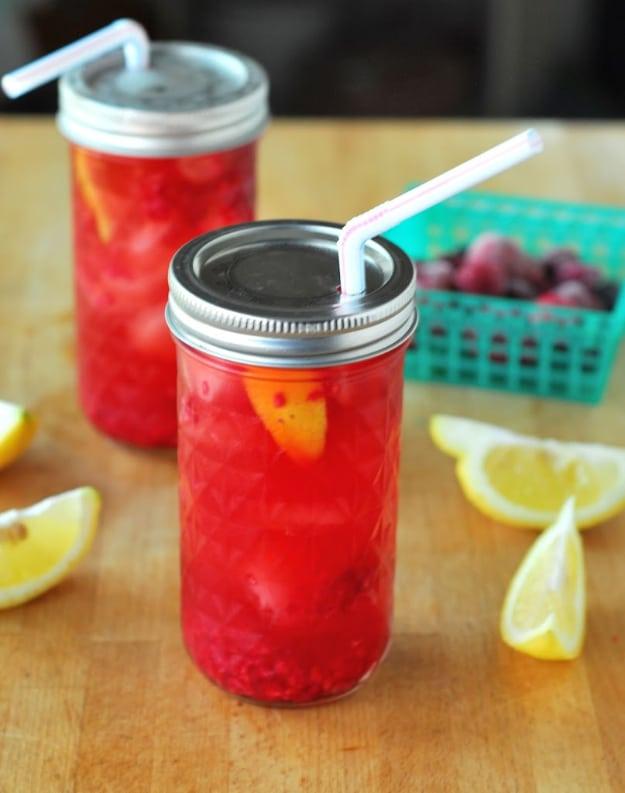 Two mason jars of red raspberry sun tea with drilled straw hole lids, lemon wedges and berries on a table