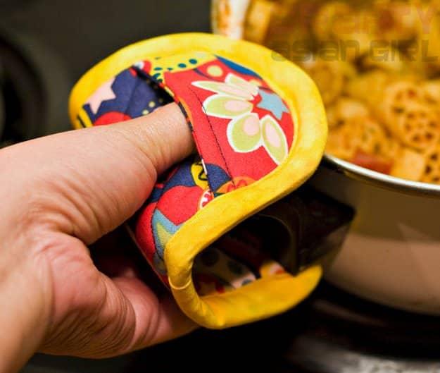 Hand holding a bright floral finger hot pad with yellow binding, gripping a saucepan handle over a stove with pasta cooking