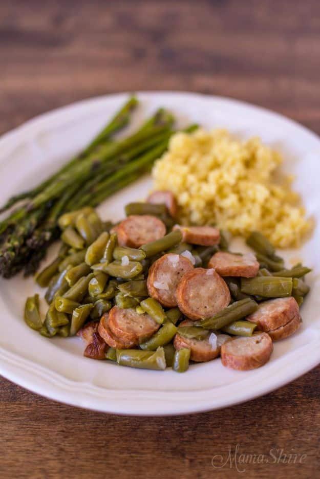 Plate of green beans with sliced sausage rounds beside asparagus and a scoop of rice on a wooden table