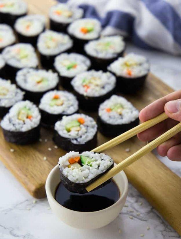 Vegan sushi rolls on a wooden board with chopsticks lifting one piece toward a small bowl of soy sauce