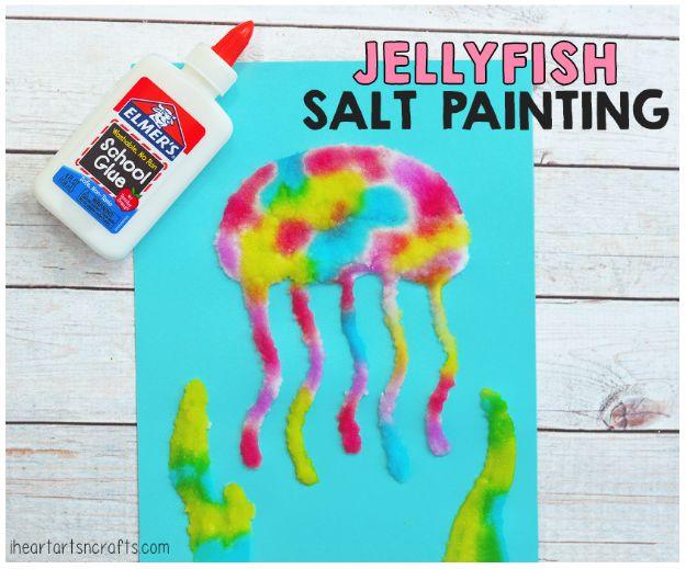 Pink and yellow jellyfish salt painting on blue cardstock with trailing tentacles, beside a school glue bottle