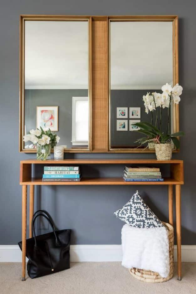 Long low wood console table with open shelf on four round tapered legs under two gold framed mirrors on a grey wall