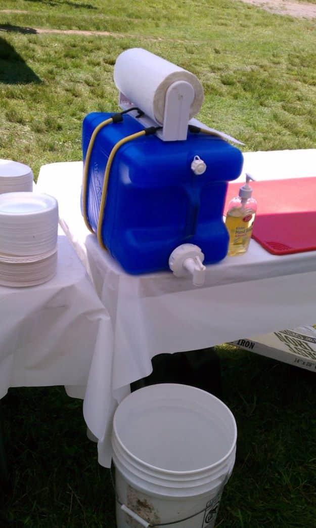 Blue water jug on a white clothed table with a paper towel roll bungeed on top, a soap bottle and a catch bucket