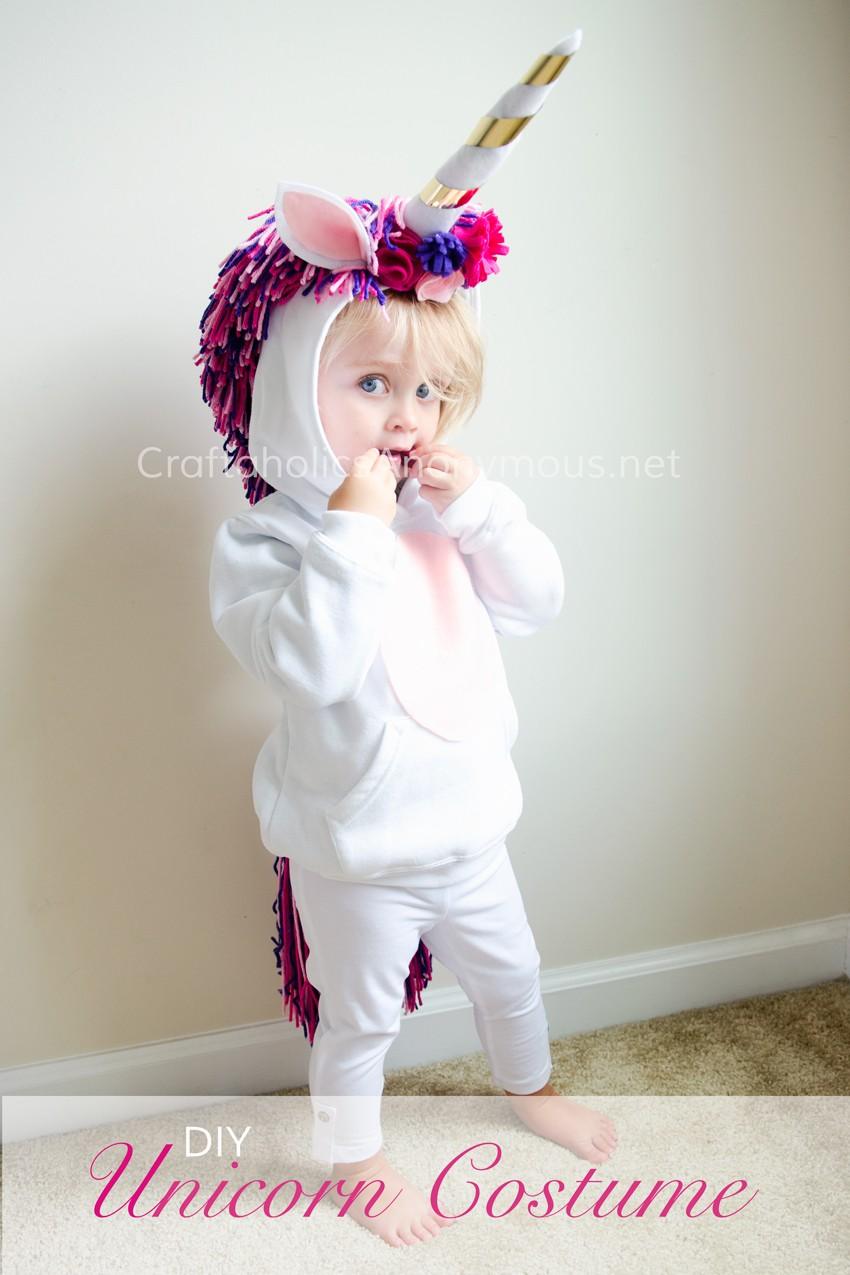 Child wearing a white unicorn hoodie costume with a gold-striped horn, felt ears, flowers and a pink-purple yarn mane