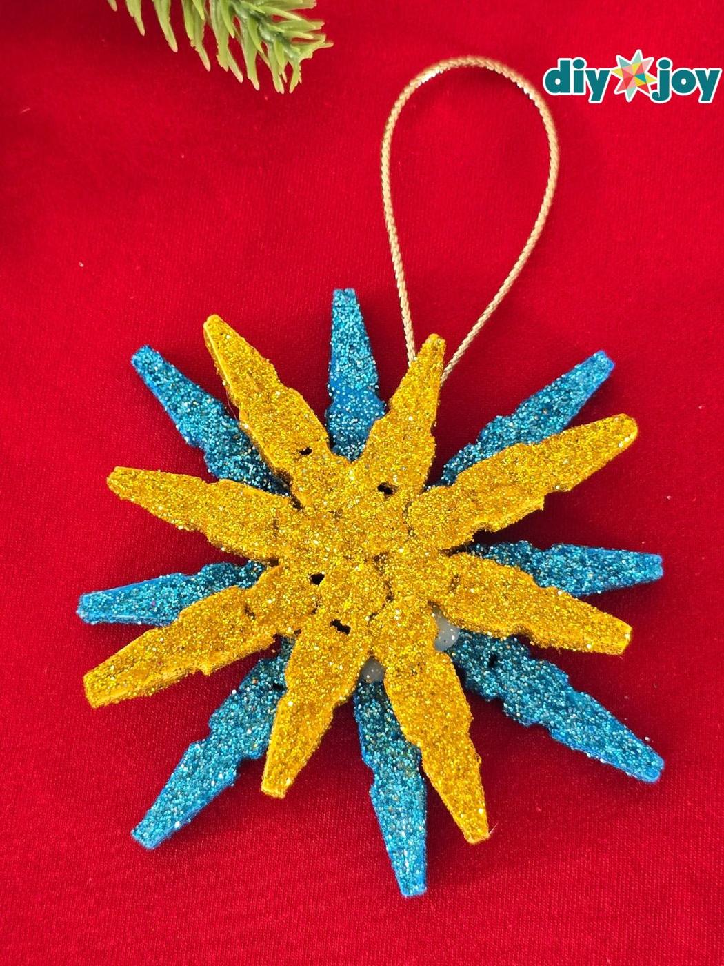 Snowflake ornament made from glittered clothespin halves in gold layered over blue, with a gold cord loop on red fabric