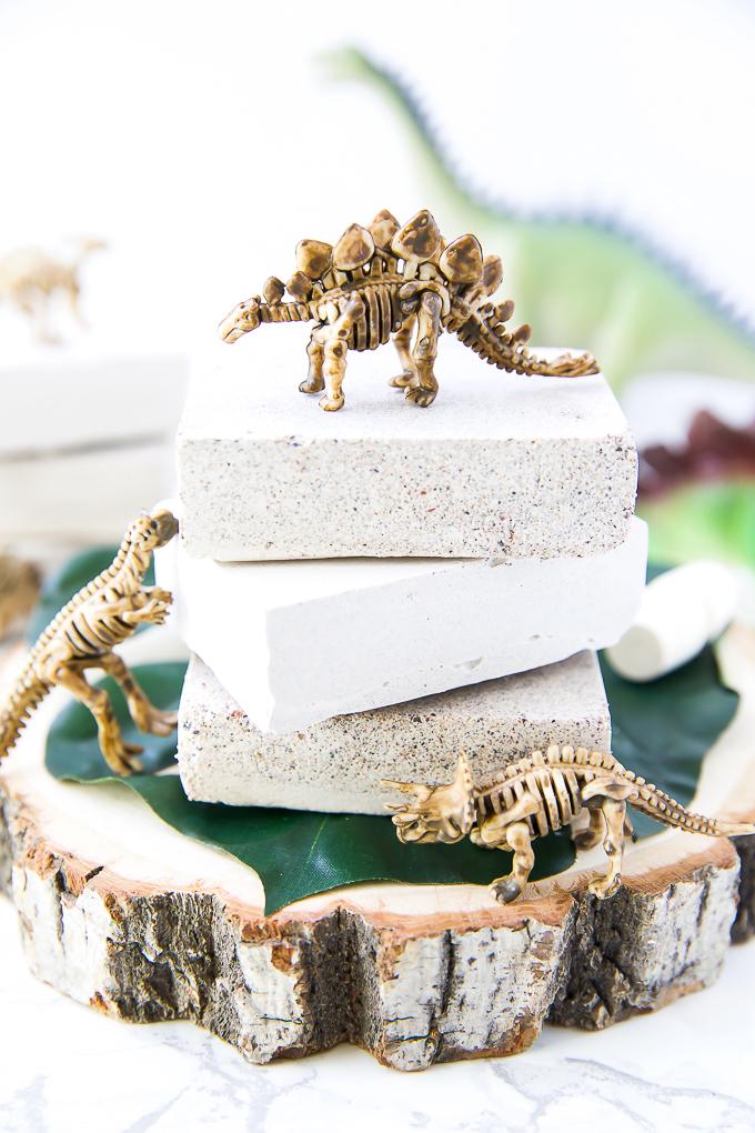 Three sandy plaster excavation bricks stacked on a wood slice with small toy dinosaur skeletons standing on and around them