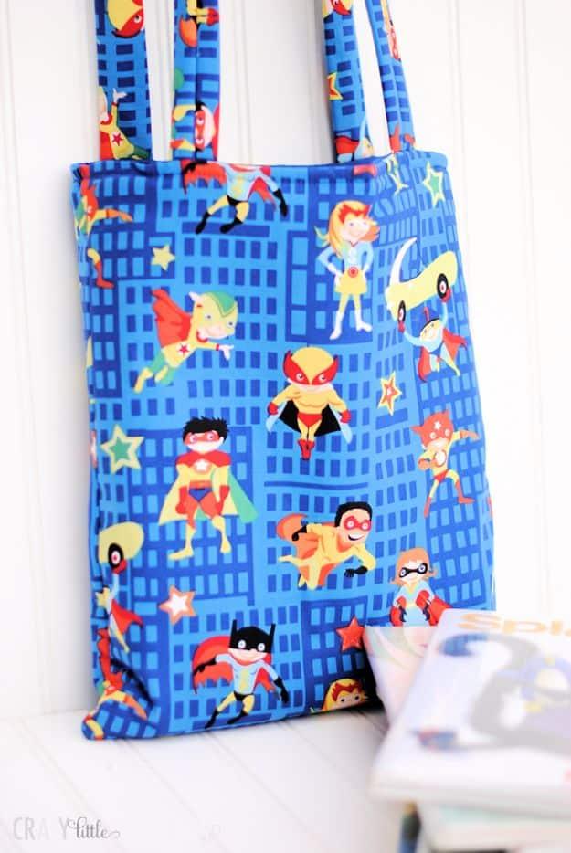 Library tote bag sewn in bright blue superhero print cotton with two shoulder straps, standing beside a stack of books