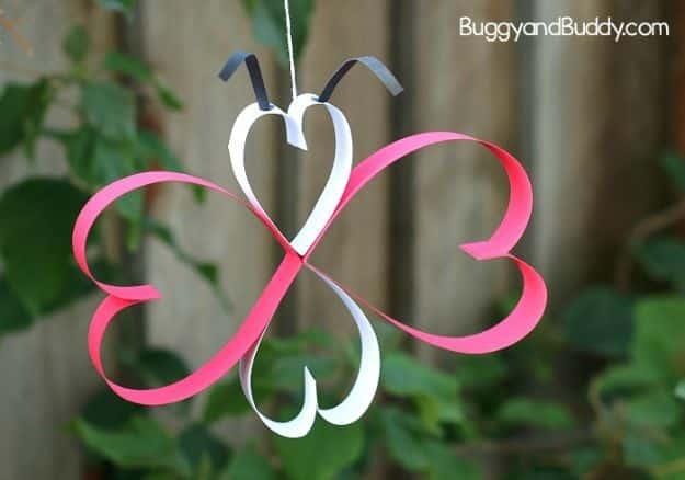 Pink and white paper heart butterfly hanging on a thread outdoors with a garden fence blurred behind it