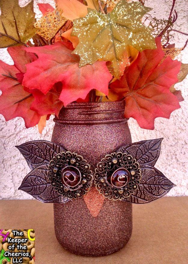 Mason jar owl covered in pink glitter spray paint with metal leaf eyes, marble pupils and fall leaves inside