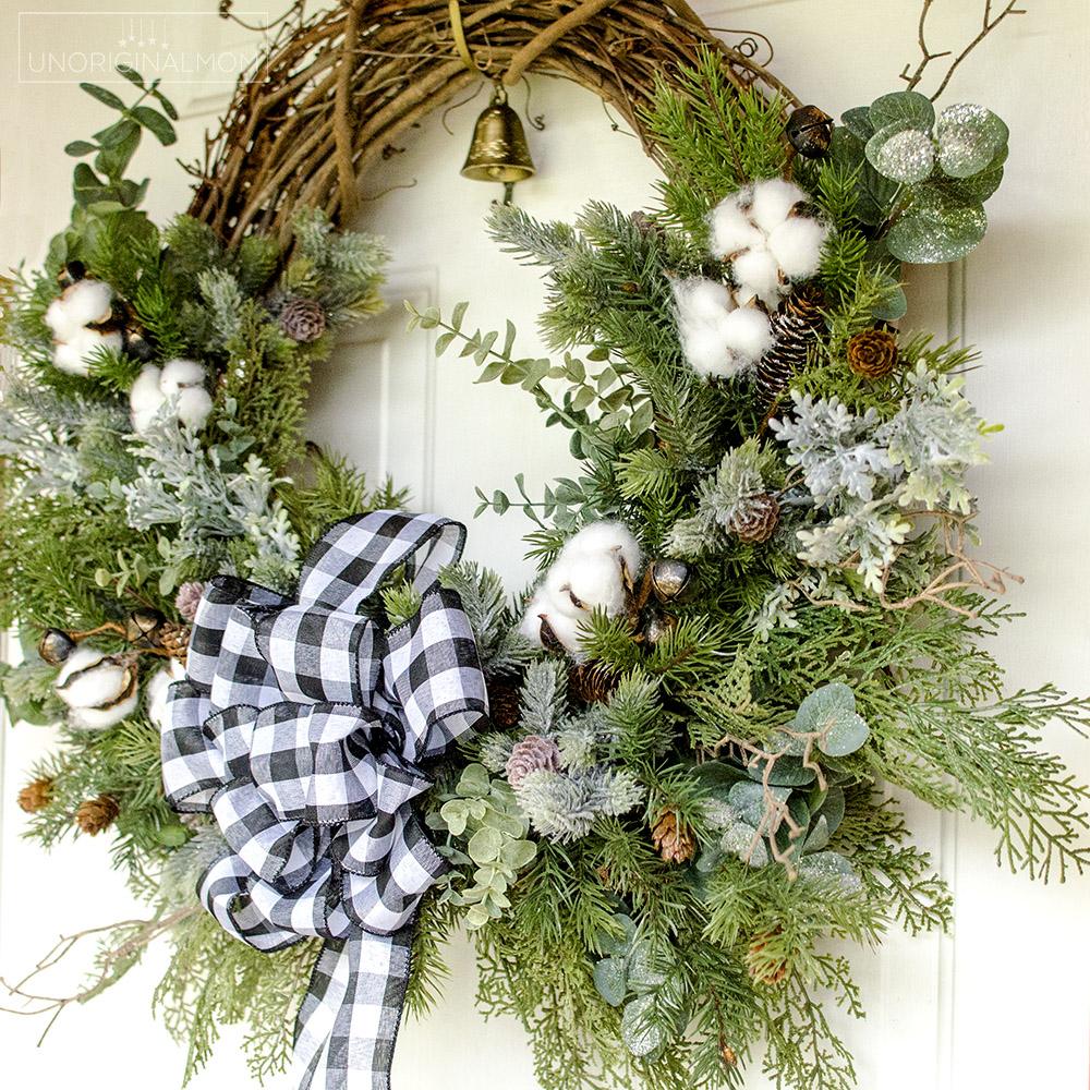 A grapevine wreath layered with flocked greenery, cotton bolls, eucalyptus, mini pinecones, bells and a buffalo check bow